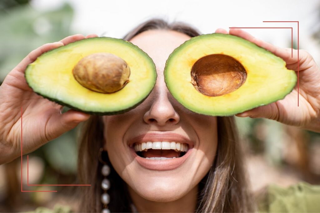 Lachende Frau hält zwei Avocadohälften vor die Augen – gesunde Ernährung mit Spaß und Leichtigkeit